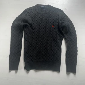 Svart kabelstickad tröja från Ralph Lauren - Kabelstickad svart tröja från Ralph Lauren med rund halsringning och klassisk röd logga broderad på bröstet. Långärmad modell i mjukt material som passar perfekt till lager-på-lager-stil. Den är i storlek m/s