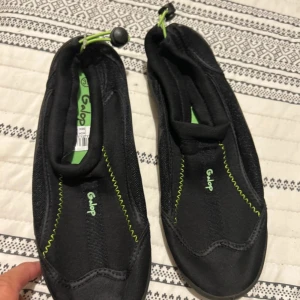 Svarta badskor från Galop, storlek 39 - Svarta badskor från Galop med gröna detaljer och synliga sömmar. Skorna har dragsko med snörlås för enkel justering och sitter tajt på foten. Perfekta för stranden eller poolen, tillverkade i syntetmaterial som torkar snabbt.
