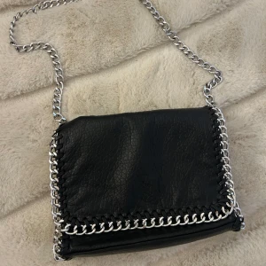 Svart axelväska med kedjedetaljer Tiamo - Svart axelväska från Tiamo i skinnimitation med grova silverfärgade kedjor längs kanterna och som axelrem. Väskan har dragkedja baktill och flera fack inuti. Perfekt för dig som gillar edgy och stilren design.