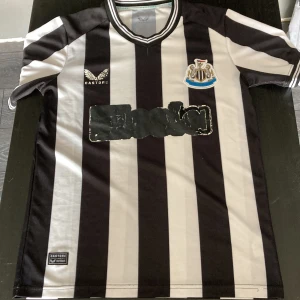 Newcastle United randig fotbollströja - Snygg Newcastle United matchtröja från Castore med klassiska svarta och vita breda ränder. Broderat klubbmärke på bröstet och Castore-logga. Tröjan har rund hals och är tillverkad i lätt, ventilerande polyester. Perfekt för dig som vill visa ditt stöd på läktaren eller hemma.