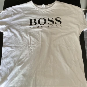 Vit t-shirt från Hugo Boss - Snygg vit t-shirt från Hugo Boss med stor svart BOSS-logga på bröstet. Klassisk rund halsringning och korta ärmar. Perfekt för dig som gillar stilrena och trendiga plagg med tydlig branding.