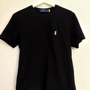 Svart t-shirt från Polo Ralph Lauren - Svart t-shirt från Polo Ralph Lauren med klassisk rund hals och korta ärmar. Diskret vit broderad logga på bröstet ger en clean och stilren vibe. Tillverkad i mjuk bomull som känns skön mot huden. Perfekt för dig som gillar enkel och snygg design.