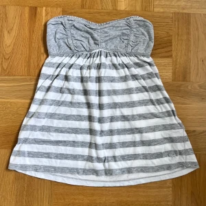 Randig vintage tubtopp från Abercrombie - Superfin tubtopp från Abercrombie & Fitch i grått och vitt med ränder. Toppen är ärmlös och har en lätt rynkad byst samt broderad älgdetalj längst ner. Perfekt för varma dagar och chill vibes. Den är vintage och säljs inte längre. Storlek M men passar XS-M skulle jag säga