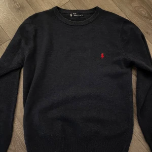  Ralph Lauren tröja - Klassisk mörk blå stickad tröja från Polo Ralph Lauren med rund halsringning och röd broderad logga på bröstet. Tröjan har lång ärm och ribbade muddar vid ärmslut och nederkant. Perfekt för lager-på-lager och snygg till jeans. priset är it hugget i sten.