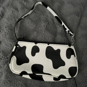 Axelväska med svartvitt ko-mönster - Säljer en axelväska i med svartvitt ko-mönster. Väskan har en smidig dragkedja upptill och ett kort axelband, perfekt för att bära det viktigaste. Den har en cool och lekfull look som sticker ut i mängden. Vid frågor hör av dig!