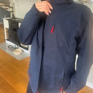 Mörkblå vindjacka Helly Hansen XL - Säljer en mörkblå vindjacka från Helly Hansen i storlek XL skulle säga passar både man och dam . Jackan har röda detaljer på dragkedjorna, hög krage och logga på ryggen och axeln. Tillverkad i polyester och har en regular passform. Perfekt för blåsiga dagar och utomhusaktiviteter.