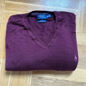 Vinröd merinoullströja Polo Ralph Lauren - Snygg vinröd stickad tröja från Polo Ralph Lauren i slim fit-modell. Tröjan är tillverkad i 100% merinoull och har en klassisk v-ringning samt den ikoniska broderade loggan på bröstet. Perfekt för dig som gillar stilrena och tidlösa plagg.