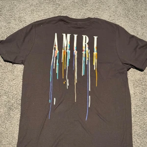 Svart AMIRI t-shirt med färgstänk - Svart t-shirt från AMIRI med coolt tryck av loggan och färgstänk i blått, gult, orange och vitt både fram och bak. Klassisk passform och rund hals. Perfekt för dig som gillar streetwear och vill sticka ut med en unik design. Aldrig använt , ny skick , ligger ute just nu för enedast 499 kr i stl M 