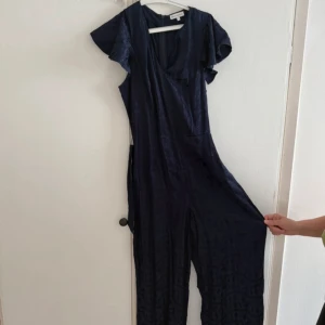 Mörkblå byxdress med v-ringning - Snygg mörkblå byxdress från warehouse med korta volangärmar och v-ringning. Byxdressen har vida ben och är tillverkad i ett lätt, glansigt tyg som ger en lyxig känsla. Perfekt för dig som gillar en avslappnad men trendig look.