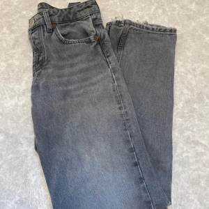 Gråa jeans straight fit - Säljer ett par gråa jeans med klassisk rak passform och fem fickor. Jeansen har hög midja, knappar i gylfen och råa benslut för en avslappnad vibe. Perfekta för dig som gillar vintagekänsla och tidlös stil.