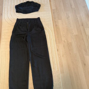 Svarta randiga kostymbyxor wide med topp - Snygga svarta kostymbyxor med vita smala ränder och wide fit. Byxorna har hög midja och klassisk kostymstil, perfekta för en trendig och stilren look. Passar dig som gillar att sticka ut med en cool och modern vibe.