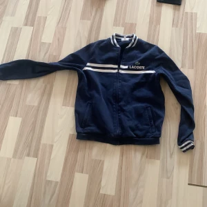 Blå bomberjacka från Lacoste Sport - Säljer en snygg mörkblå bomberjacka från Lacoste Sport med vita ränder på bröstet och kragen. Jackan har dragkedja, ribbade muddar och två fickor framtill. Perfekt för dig som gillar sportig och stilren look.