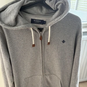 Grå hoodie från Morris Company - Snygg grå hoodie från Morris Company med dragkedja, huva och vita snören. Tröjan har en liten broderad logga på bröstet och praktiska fickor framtill. Har inte kommit till användning på länge, inga speciella skäl. Den har inga alls defekter. Skriv vid minsta fråga!