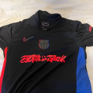 FC Barcelona x Nike svart fotbollströja - Snygg svart FC Barcelona fotbollströja från Nike med röda och blå detaljer på sidorna. Tryck med Nike-logga, FCB-märke och Cactus Jack-grafik på bröstet. UNHCR-logga på ryggen. Tillverkad i Dri-Fit-material som andas och håller dig torr. Storlek S men passar även M, använd en gång bara inga defekter alls. Pris kan diskuteras vid snabb affär.
