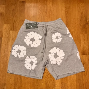 Grå shorts med blommor från Denim Tears - Grå shorts från Denim Tears med stora vita blommiga tryck över hela plagget. Modellen har elastisk midja med snörning för skön passform och är tillverkad i mjuk bomull. Perfekt för chill dagar och streetwear vibes.