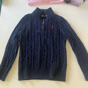 Marinblå kabelstickad tröja Ralph Lauren - Kabelstickad marinblå tröja från Polo Ralph Lauren med halv dragkedja och hög krage. Tröjan har lång ärm, ribbade muddar och röd broderad logga på bröstet. Perfekt för dig som gillar klassisk och stilren look. Pris kan diskuteras. Skriv för fler bilder 