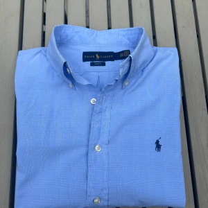 Ljus blå Ralph Lauren skjorta i M/36 37 - Klassisk ljusblå skjorta från Ralph Lauren i slim fit-modell. Skjortan är väldigt classy och lyxig. Den har en svart logga och är tillverkad i bommul.  (Jag tror den är i m)