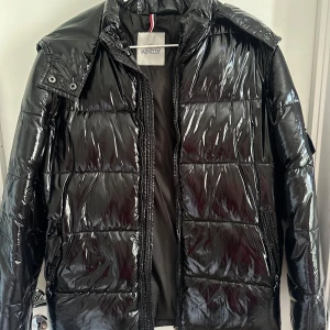 Svart glansig pufferjacka från Moncler - Säljer en svart, glansig pufferjacka från Moncler med quiltad design och dragkedja framtill. Jackan har hög krage och avtagbar huva, perfekt för kalla dagar. Klassisk och stilren look som lyfter vilken outfit som helst.