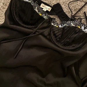 Svart miniklänning i satin med broderier  - Super charmig svart miniklänning i satin från For Love Bell med tunna spagettiband, insydda kupor och spetskant med broderade blommor i lila, grönt och gult. Söt rosett framtill och öppen rygg med cool cut-out detalj. Perfekt för dig som gillar en chic och feminin look. 😋✨🖤🩷Använd endast en gång! Storlek M men passar även en S.