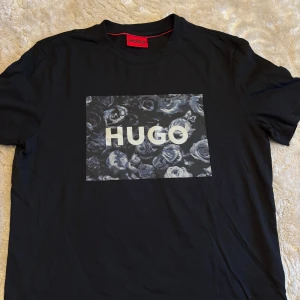 Svart HUGO t-shirt med blommigt tryck - Svart  t-shirt från HUGO i bomull med ett stort svart blommigt tryck och HUGO-logga på bröstet. T shirten är i storlek S och helt oanvänd. Köpt men bara legat i garderoben derför skrynklig annars är den helt ny. Hör av vid frågor. 