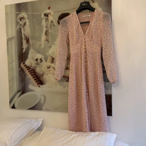 Ljusrosa blommig långklänning Adoore - Supersöt långklänning från Adoore i ljusrosa med småblommigt mönster. Klänningen har v-ringning, långa ärmar och ett flowy fall. Perfekt för vår och sommar, och ger en romantisk vibe med sina fina detaljer.