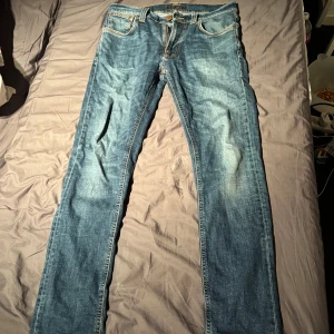 Blå jeans från Nudie Jeans - Säljer ett par klassiska blå jeans från Nudie Jeans med snyggt slitna detaljer och kontrastsömmar. Modellen har raka ben och fem fickor, samt Nudie Jeans signaturbroderi på bakfickorna. Perfekta för dig som gillar en tidlös och avslappnad look.