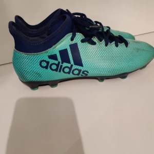 Adidas Techfit X blågröna fotbollsskor - Säljer ett par Adidas Techfit X fotbollsskor i blågrönt med mörkblå detaljer och snörning. Skorna har en prickig design och är tillverkade i syntetmaterial. Perfekta för dig som vill sticka ut på planen med stilrena och sportiga skor.