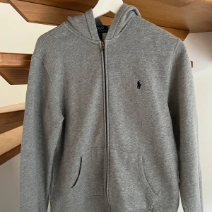 Grå hoodie från Ralph Lauren - Säljer en klassisk grå hoodie från Ralph Lauren med dragkedja och fickor framtill. Tröjan har en liten broderad logga på bröstet och är perfekt för en avslappnad stil. Mjuk och bekväm med långa ärmar och huva. Ordinarie pris 1195 kr. 