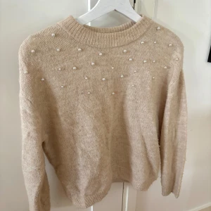 Beige stickad tröja med pärlor - Superfin beige stickad tröja med pärldetaljer upptill. Tröjan har rund halsringning och långa ärmar, perfekt för att ge en söt och trendig touch till din outfit. Mjuk och skön att bära under kyligare dagar nu till hösten!