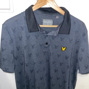 Blå Lyle & Scott t-shirt med krage - Snygg blå t-shirt från Lyle & Scott med klassisk pikékrage och korta ärmar. T-shirten har ett subtilt mönster med små fåglar över hela tyget och en gul broderad logga på bröstet. Perfekt för dig som gillar stilrena detaljer och skön passform.