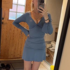 Blå klänning med snörning - Supersöt blå kortklänning med v-ringning. Långa ärmar med snörning vid handlederna och öppen rygg med knytband. Sitter figurnära och har lätt utsvängd kjol. Perfekt för dig som gillar trendiga detaljer och en feminin look.