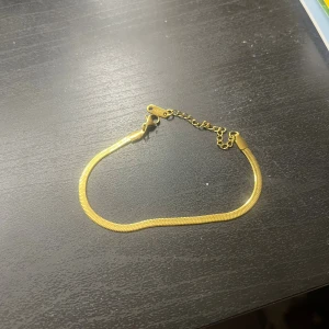 Guldigt kedjearmband med justerbar längd - I rostfritt stål. Snyggt och stilrent armband i guldfärgad metall med en platt kedja och justerbar längd. Armbandet har en smidig karbinlås och extra kedja för att passa olika handleder. Perfekt för att ge din look lite extra shine.