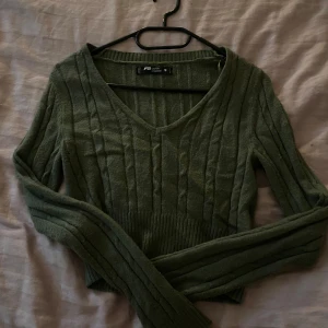 Olivgrön stickad tröja från FB Sister - Snygg olivgrön stickad tröja från FB Sister med v-ringning och ribbade detaljer i midjan. Tröjan har lång ärm och ett ribbat, croppat avslut som ger en modern look. Perfekt att styla med jeans eller kjol för en chill vibe.