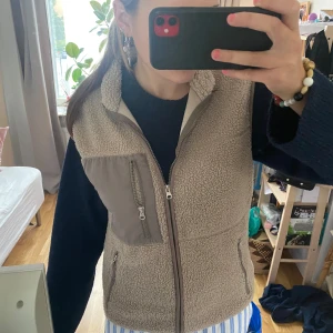 Teddyjacka väst  - Mysig beige fleeceväst från 157 med hög krage och tre fickor med dragkedja, varav en bröstficka i kontrasterande tyg. Perfekt att slänga över en hoodie för extra värme. Västen har en rak passform och är superenkel att styla till vardags.