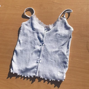 Vit ribbad linnetopp från Censored - Supersöt vit ribbad linnetopp från Censored i storlek XXS. Toppen har tunna axelband, spetskant vid urringningen och små rosetter framtill. Den är croppad med vågig kant nertill och har en mjuk, stretchig känsla. Perfekt för en chill och trendig look.