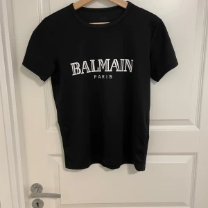 Svart Balmain Paris t-shirt - Svart t-shirt från Balmain med vit logotyptext 'BALMAIN PARIS' på bröstet. Klassisk rund hals och korta ärmar. Stilren och enkel design som passar perfekt till jeans eller shorts. Materialet känns mjukt och skönt mot huden.