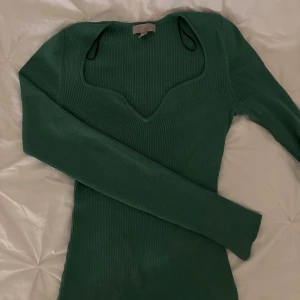 Grön ribbad tröja från H&M - Mörkgrön ribbad tröja från H&M med lång ärm och snygg v-ringad halsringning. Tröjan är figurnära och har en mjuk, stretchig känsla som gör den superbekväm. Perfekt att styla med jeans eller kjol för en trendig look.
