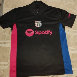 FC Barcelona svart fotbollströja - Säljer en FC Barcelona fotbollströja i svart med rosa och blå sidopaneler. Tröjan har korta ärmar, klubbmärke på bröstet och Spotify-logga framtill. Baksidan har UNHCR ACNUR-tryck. Tillverkad i lätt polyester, perfekt för match eller träning.