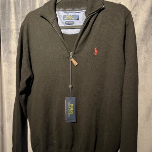 Svart Polo Ralph Lauren halv zip - Svart långärmad tröja från Polo Ralph Lauren i mjuk pima bomull. Klassisk half zip med läderdetalj på dragkedjan och röd broderad logga på bröstet. Perfekt för dig som gillar stilren och tidlös design. Knappt använd säljes i nyskick