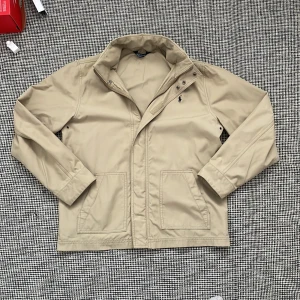 Beige vindjacka/fieldjacka från Polo Ralph Lauren -  beige vindjacka/fieldjacka från Polo Ralph Lauren med mörkblå broderad logga på bröstet. två stora fickor framtill och stängs med dragkedja och tryckknappar. Perfekt för dig som gillar stilren design. Passar perfekt nu till hösten.