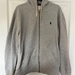 Grå hoodie från Polo Ralph Lauren - Klassisk grå hoodie från Polo Ralph Lauren med dragkedja framtill, huva med snörning och ficka. Liten broderad logga på bröstet. Storlek L