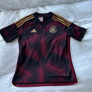 Tysklands matchtröja Adidas svart/röd - Snygg Tysklands fotbollströja från Adidas med svart bas och mörkröda grafiska mönster. Guldiga detaljer på axlar och logga, samt broderat landslagsemblem med fyra stjärnor. Kortärmad och tillverkad i lätt, ventilerande polyester. Perfekt för dig som älskar fotboll och vill sticka ut.