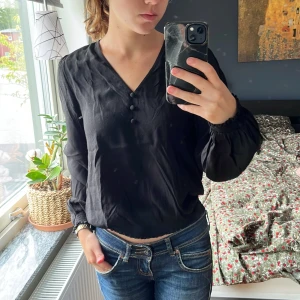 Svart blus med v-ringning från EDC - Svart blus från EDC med v-ringad hals och tre dekorativa knappar framtill. Långa ärmar med smock vid mudden och lätt rynkad nederkant. Materialet är mjukt och har ett snyggt fall, perfekt till jeans eller kjol för en chill look.