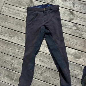 Mörkblå ridbyxor Jackie stl 152 - Snygga mörkblå ridbyxor från Jackie i stretchigt bomullsmaterial med förstärkning på insidan av benen och sätet. Byxorna har bälteshällor, dragkedjeficka och knäppning fram. 