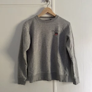 Grå sweatshirt från GANT - Säljer en stilren grå sweatshirt från GANT med broderad logga och sköld på bröstet. Tröjan har rund hals, långa ärmar och ribbade muddar. Skriv för vidare info