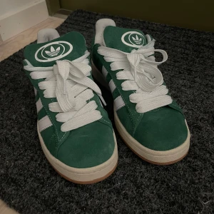 Adidas Campus gröna  - Säljer ett par gröna Adidas Campus sneakers i mocka med klassiska vita ränder och vit sula. Skorna har rund tå, platt gummisula och breda vita snören. Perfekta för dig som gillar retrostil och vill sticka ut med färg. Adidas-logga på tungan och sidan. Tillkommer gröna skosnören. Använda två gånger så i nyskick. Pris kan diskuteras vid snabb affär.