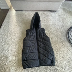 Svart dunväst med huva från Name It - Svart quiltad dunväst från Name It med huva och dragkedja framtill. Västen har en stilren look med horisontella sömmar och en reflekterande detalj baktill. Perfekt för lager-på-lager under kyliga dagar. Har använt den en gång.