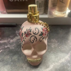 Police To Be Tattooart parfym 40ml - Unik parfymflaska från Police i form av en ljusrosa dödskalle med tatueringsinspirerade rosor och svarta mönster. Flaskan har guldfärgad spraydel och kedja. Rymmer 40 ml och är tillverkad i Italien. Perfekt för dig som gillar edgy och snygg design.