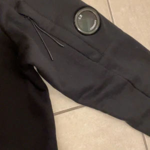 Svart sweatshirt från C.P. Company - Svart sweatshirt från C.P. Company med klassisk rund hals och lång ärm. Ärmen har en ikonisk linsdetalj och en liten ficka med dragkedja. Tröjan är i mjuk bomull och har en clean, stilren look som passar perfekt till streetwear.