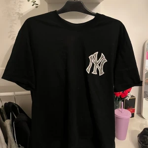 Svart New Era Yankees t-shirt - Svart t-shirt från New Era med stor New York Yankees-logga i silver på ryggen och mindre logga på bröstet. Klassisk rund hals och korta ärmar. Materialet är mjuk bomull, perfekt för dig som gillar sportiga och street-inspirerade plagg.